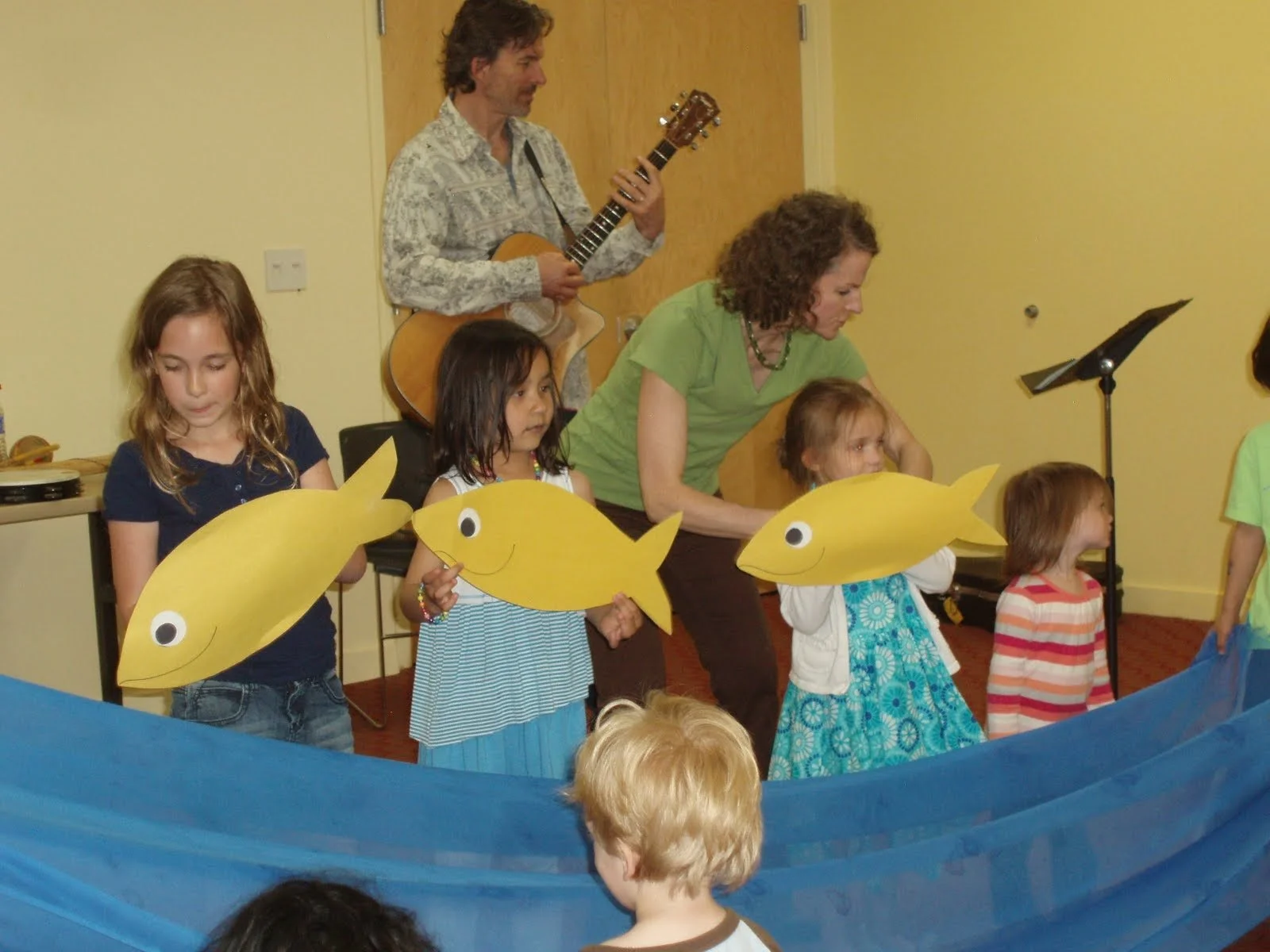 A group of children and adults performing a puppet show with yellow fish puppets, while a man in the background plays guitar, in a room with yellow walls.