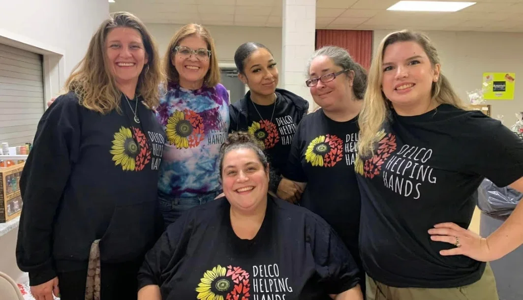 Group of six women smiling, wearing matching black T-shirts with a sunflower and red floral design and the words "Delco Helping Hands." They are indoors in a room with a table on the left and a yellow sign in the background.
