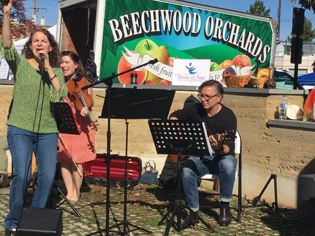 Musicians performing at Beechwood Orchards during a daytime outdoor event, with a woman singing into a microphone, a woman playing a violin, and a man playing guitar, in front of a colorful banner advertising fresh fruit.