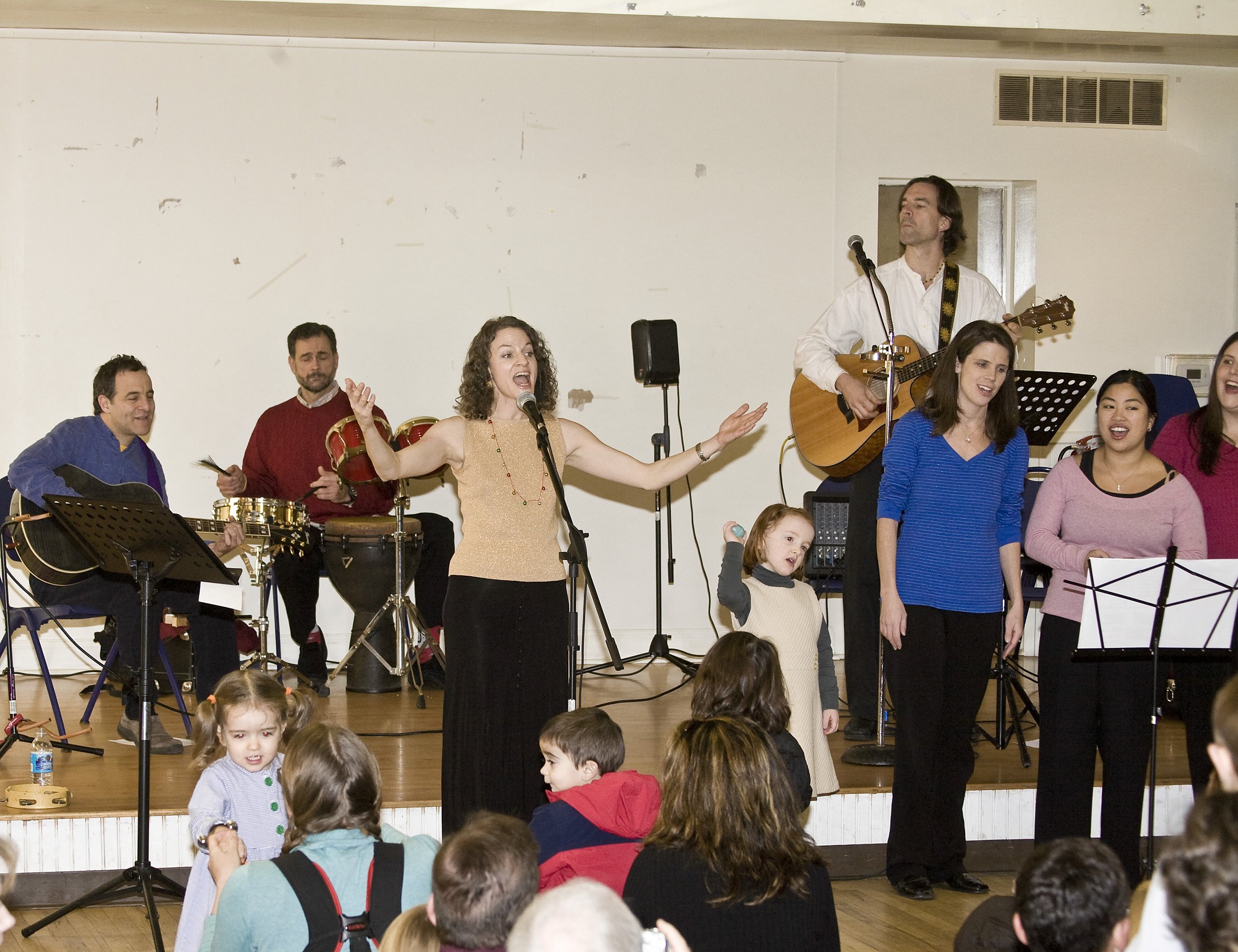 A group of people performing on a stage with children and an audience in front, singing and playing instruments.