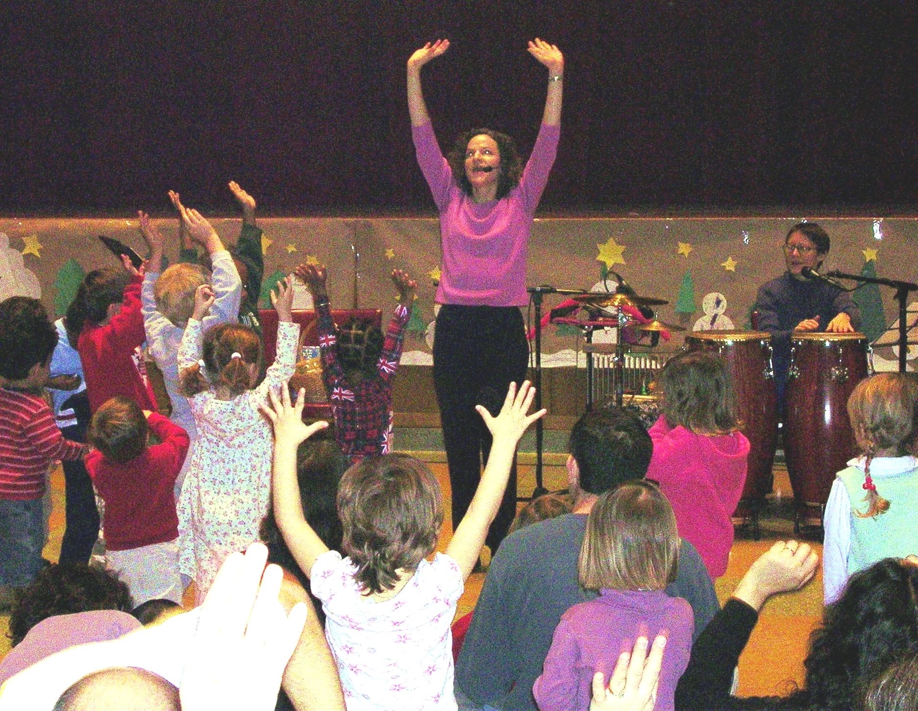 A woman in a pink top leading a group of children in a dance or musical activity in a decorated indoor space.