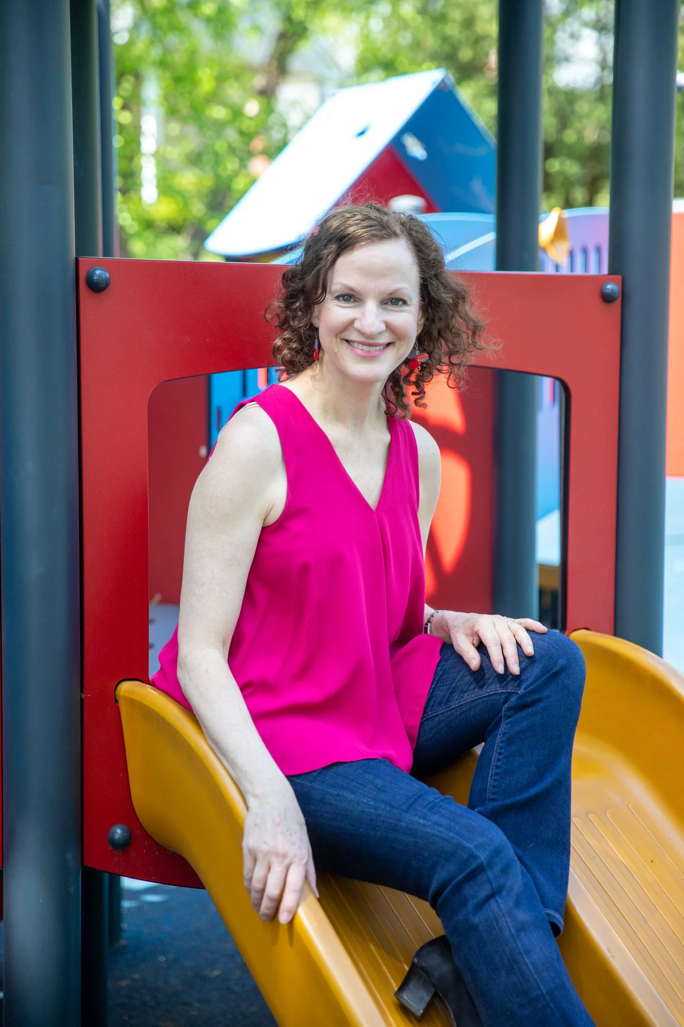 A woman with curly brown hair smiling while sitting at the top of a yellow slide in a playground, wearing a bright pink sleeveless top and dark jeans.