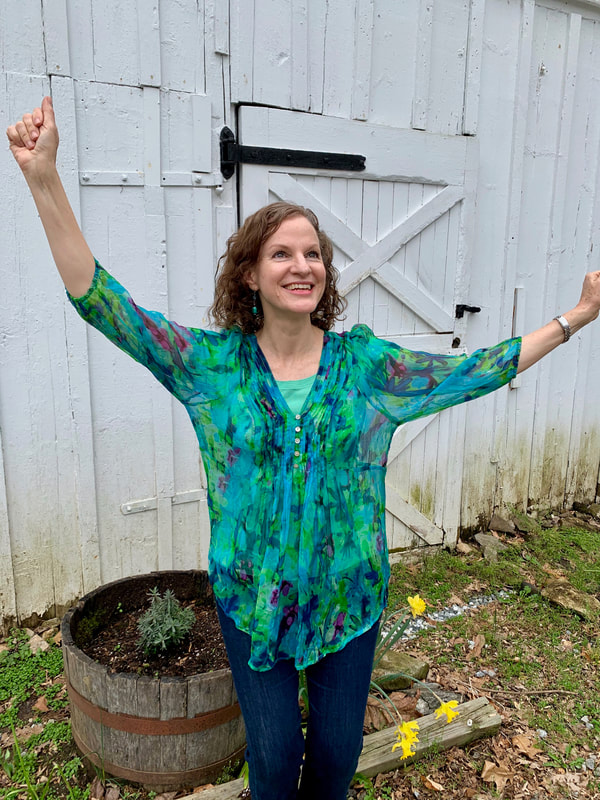 A woman with curly hair wearing a colorful blue and green blouse and jeans, smiling and raising her arms in front of a white wooden shed with a black latch and a potted plant with yellow flowers nearby.