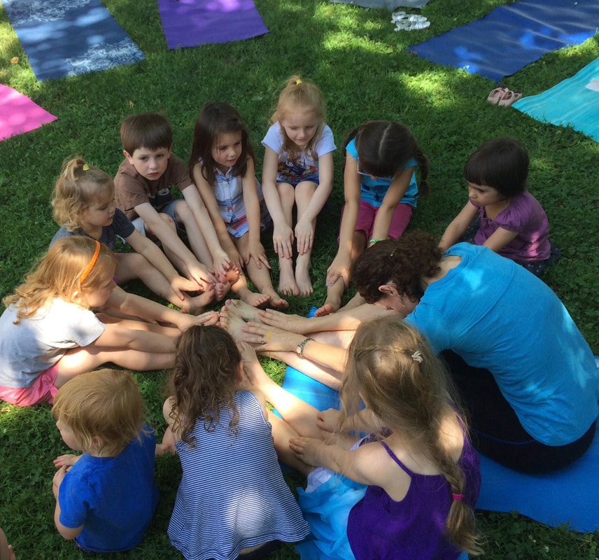 A group of children and an adult girl sitting in a circle on grass, engaging in a group activity; all touching a blue mat in the center. Outdoor setting with colorful mats laid out on the grass.