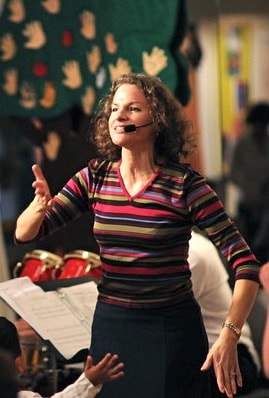 A woman with curly hair, wearing a striped long-sleeve shirt, speaking into a headset microphone in a lively classroom or workshop setting.