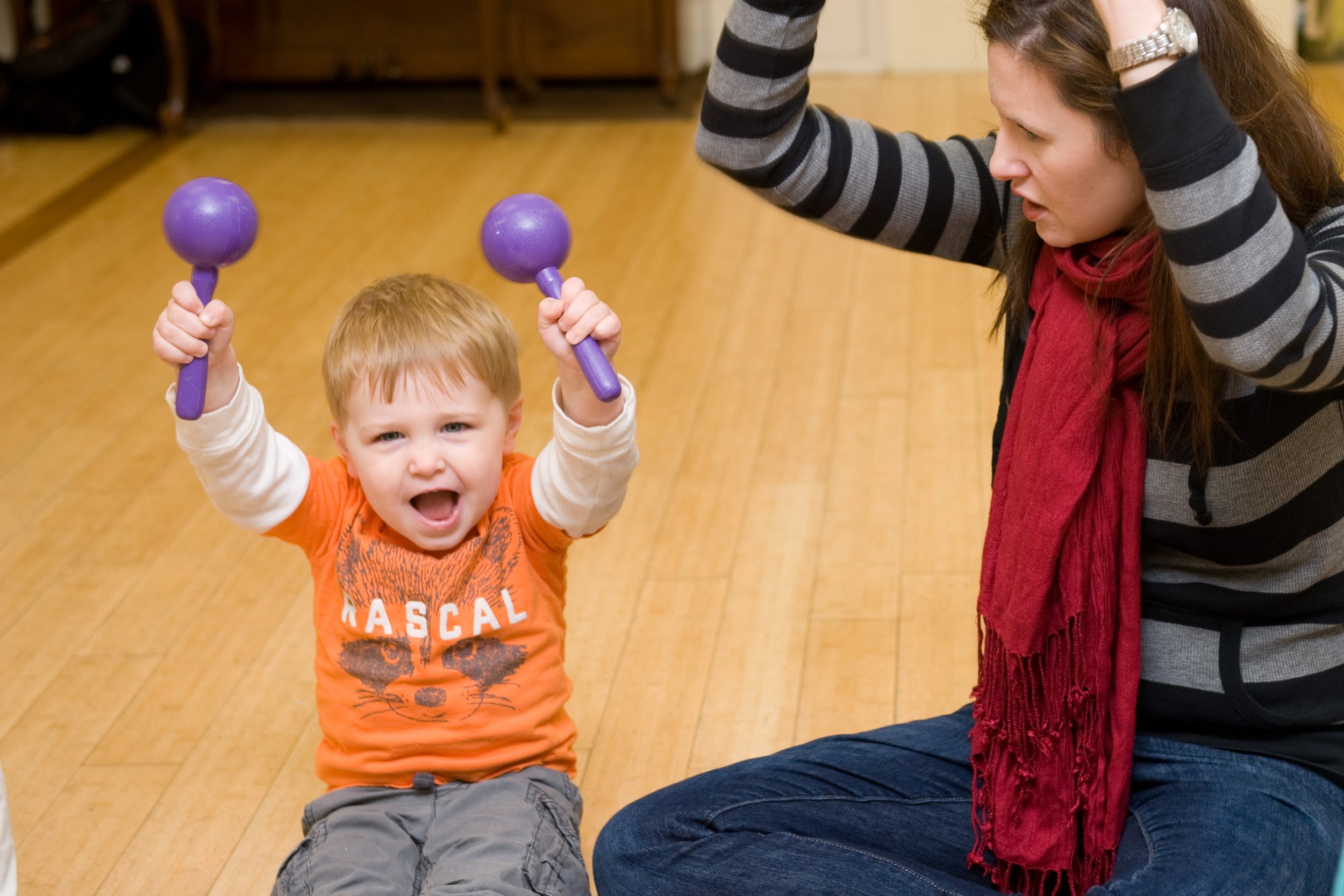 A young boy with short blonde hair and a wide open mouth, holding two purple maracas, sitting on the floor in a room with wooden flooring. A woman with long brown hair, wearing a black and grey striped sweater and red scarf, is sitting next to him, raising her hands above her head, engaging with the boy.