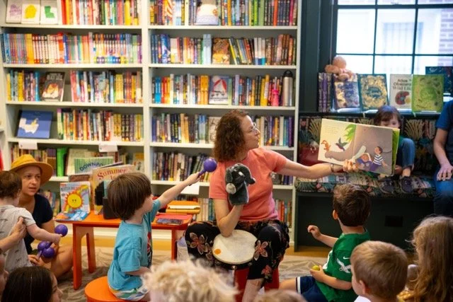A woman reading a children's book to a group of kids in a library, with bookshelves in the background.
