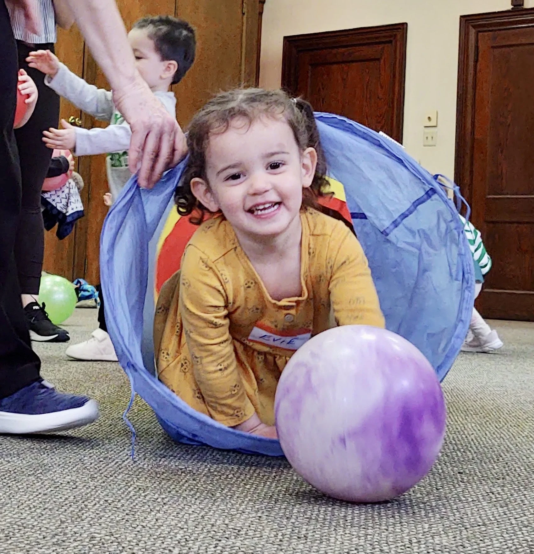 A young boy with curly brown hair and a yellow shirt smiling while crawling inside a blue play tunnel, with a purple balloon nearby, during a children's birthday party.