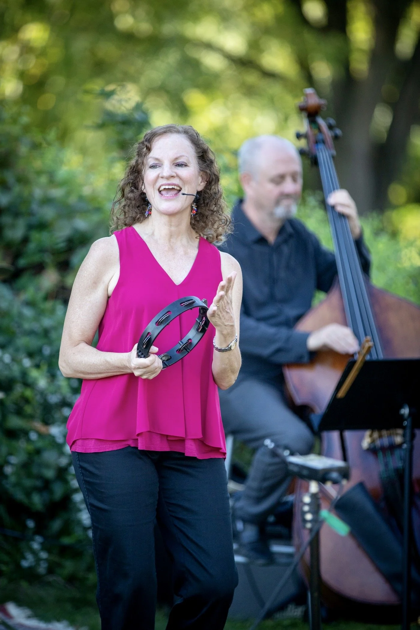 A woman with curly brown hair, wearing a bright pink sleeveless top and black pants, is playing a tambourine and singing with a smile, outdoors with trees in the background. A man with gray hair and beard, dressed in black, is playing a guitar in the background.