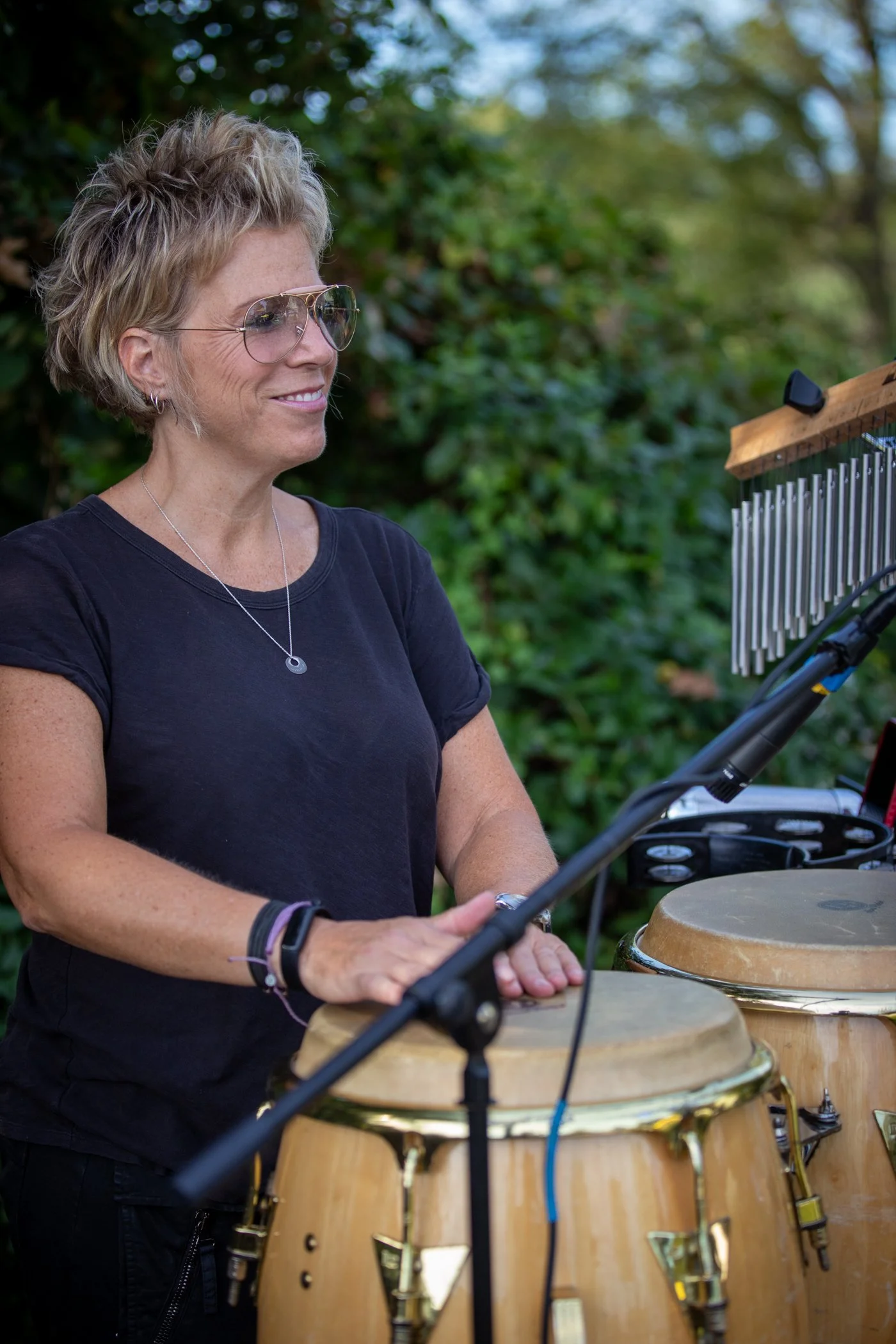 A woman with short curly blonde hair, wearing glasses and a black shirt, playing multiple hand drums outdoors, with greenery in the background.