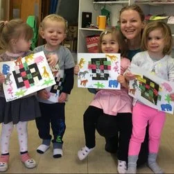 Four young girls and a woman smiling and holding up colorful pixel art projects in a classroom.