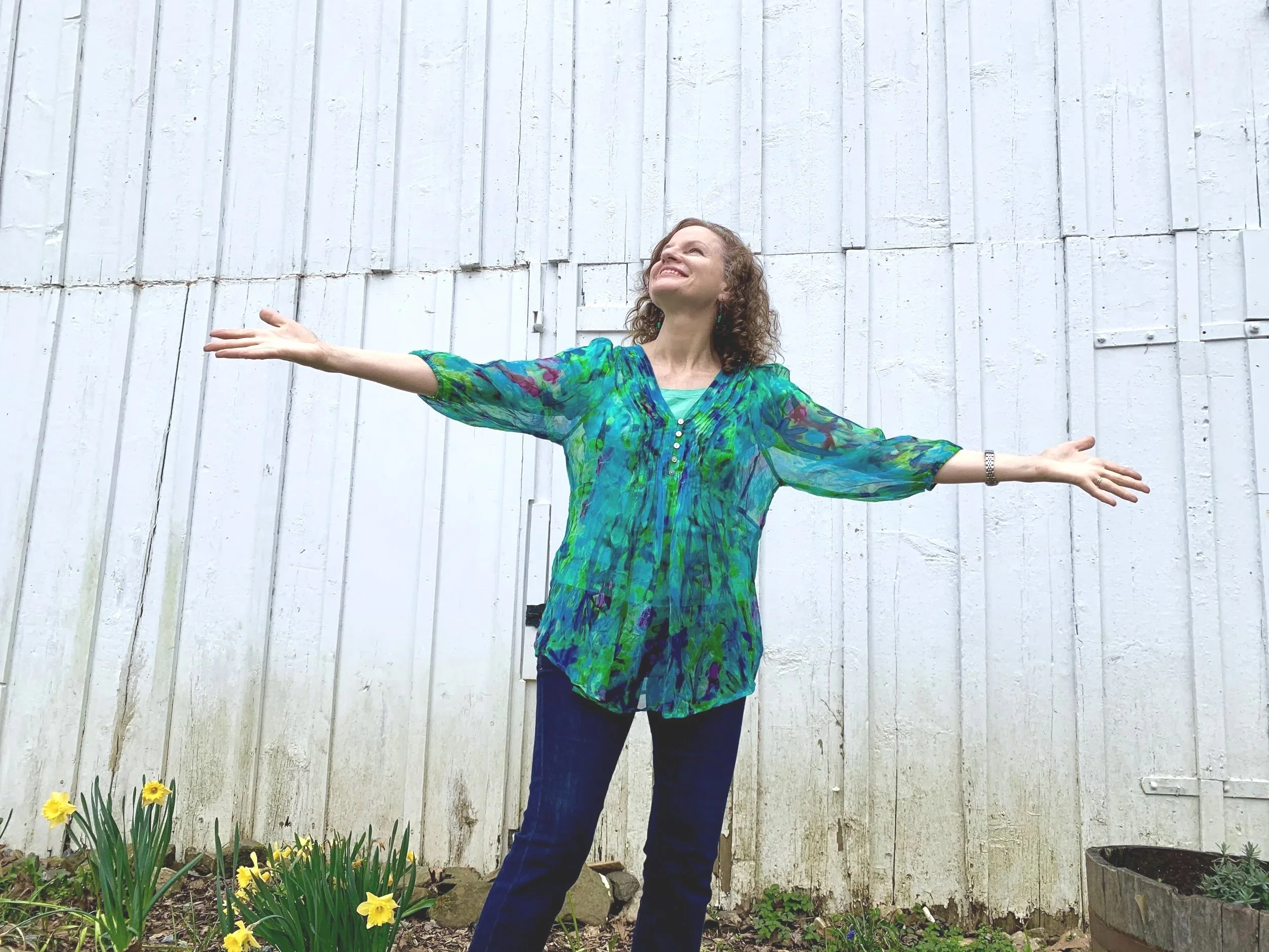 A woman with curly hair wearing a colorful green and blue patterned blouse standing outdoors with arms outstretched in front of a white wooden fence, with yellow spring flowers at her feet.