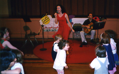 A woman singing on stage with two men playing guitars, and children in the audience watching.