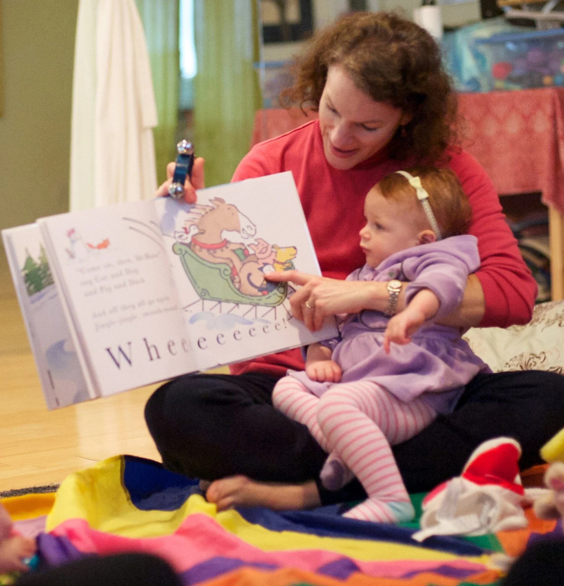 A woman reading a colorful children's book to a young girl seated on her lap, with bright multicolored paper strips on the floor in front of them.