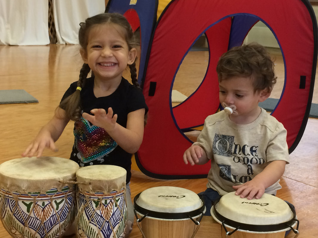 Two children playing drums indoors, a girl smiling and a boy with a pacifier, with a colorful play tunnel in the background.