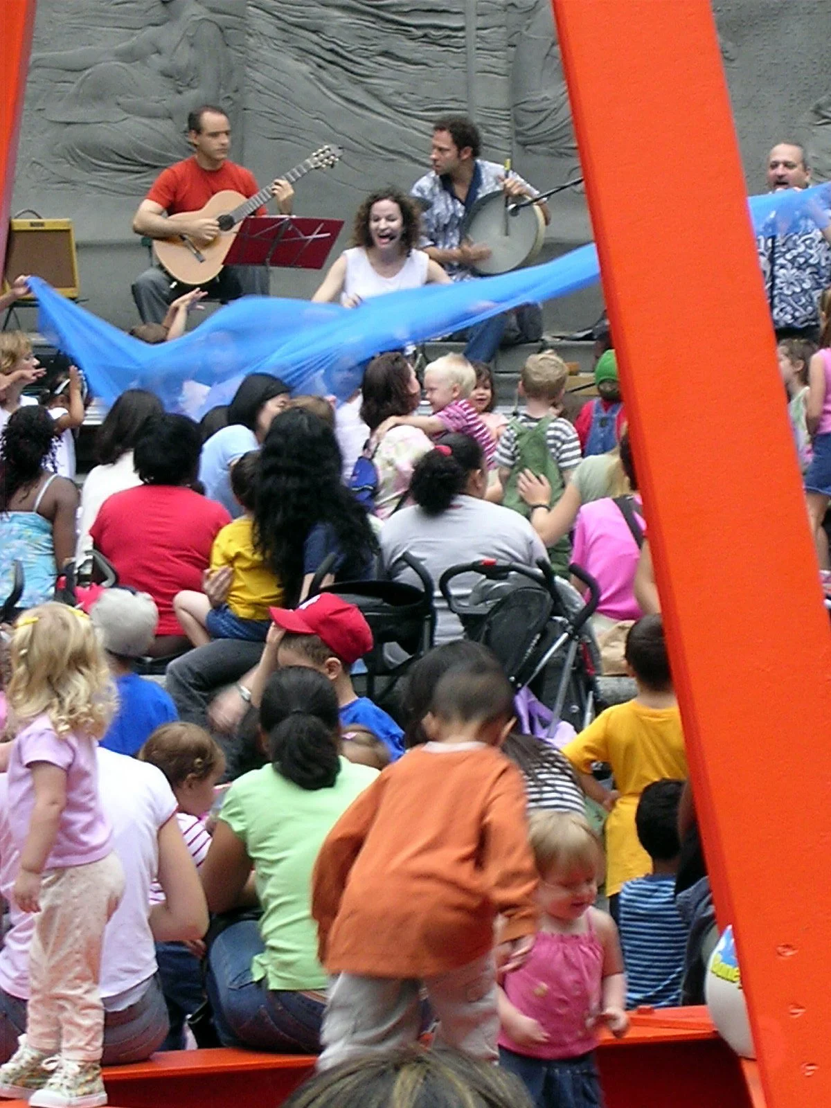 Children and adults seated on the ground in front of an outdoor stage, where three performers entertain with musical instruments and singing, with a colorful slide partially visible in the foreground.