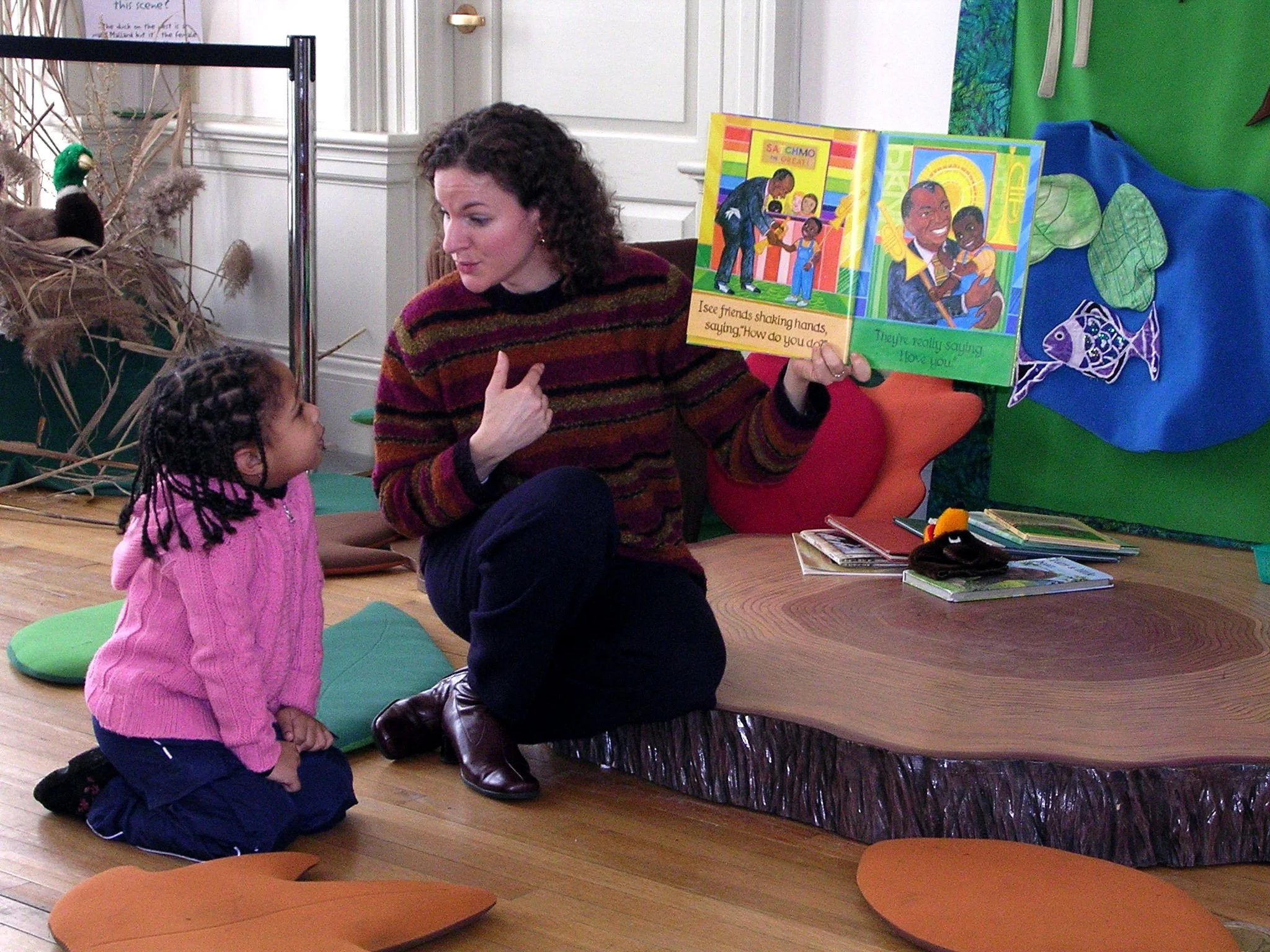 A woman reading a children's book to a young girl in a library or classroom. The woman is seated and holding the book open, showing colorful illustrations and text. The girl is kneeling on a green cushion, watching and listening attentively. There ar