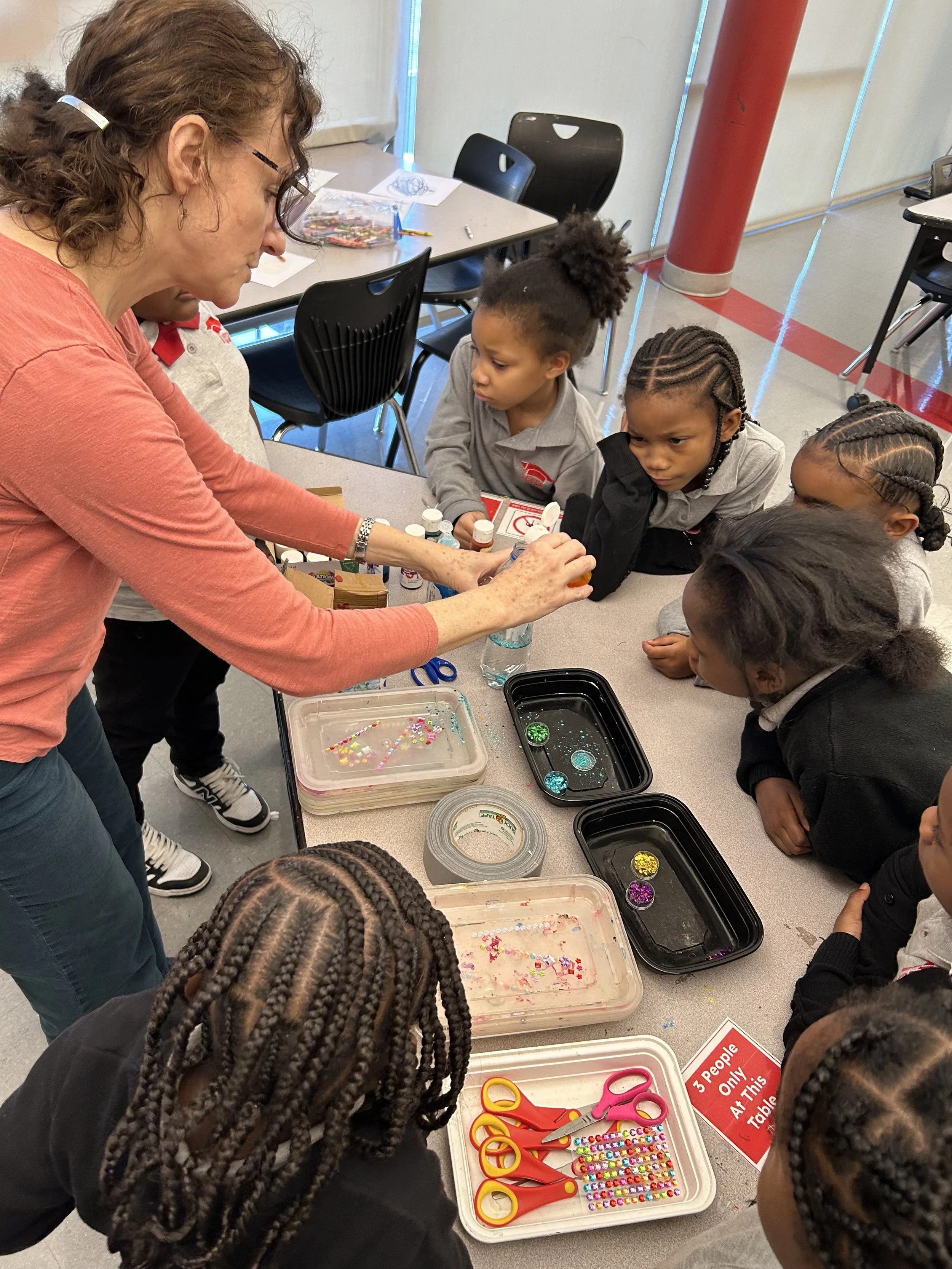 A woman demonstrating a craft activity to a group of young children, with various craft supplies such as scissors, beads, and sequins on the table.