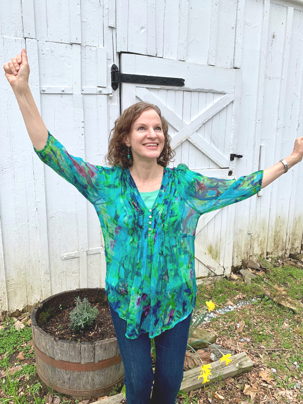 A woman with curly hair wearing a colorful green and blue top, smiling with her arms raised, standing outdoors in front of a white wooden fence and door.