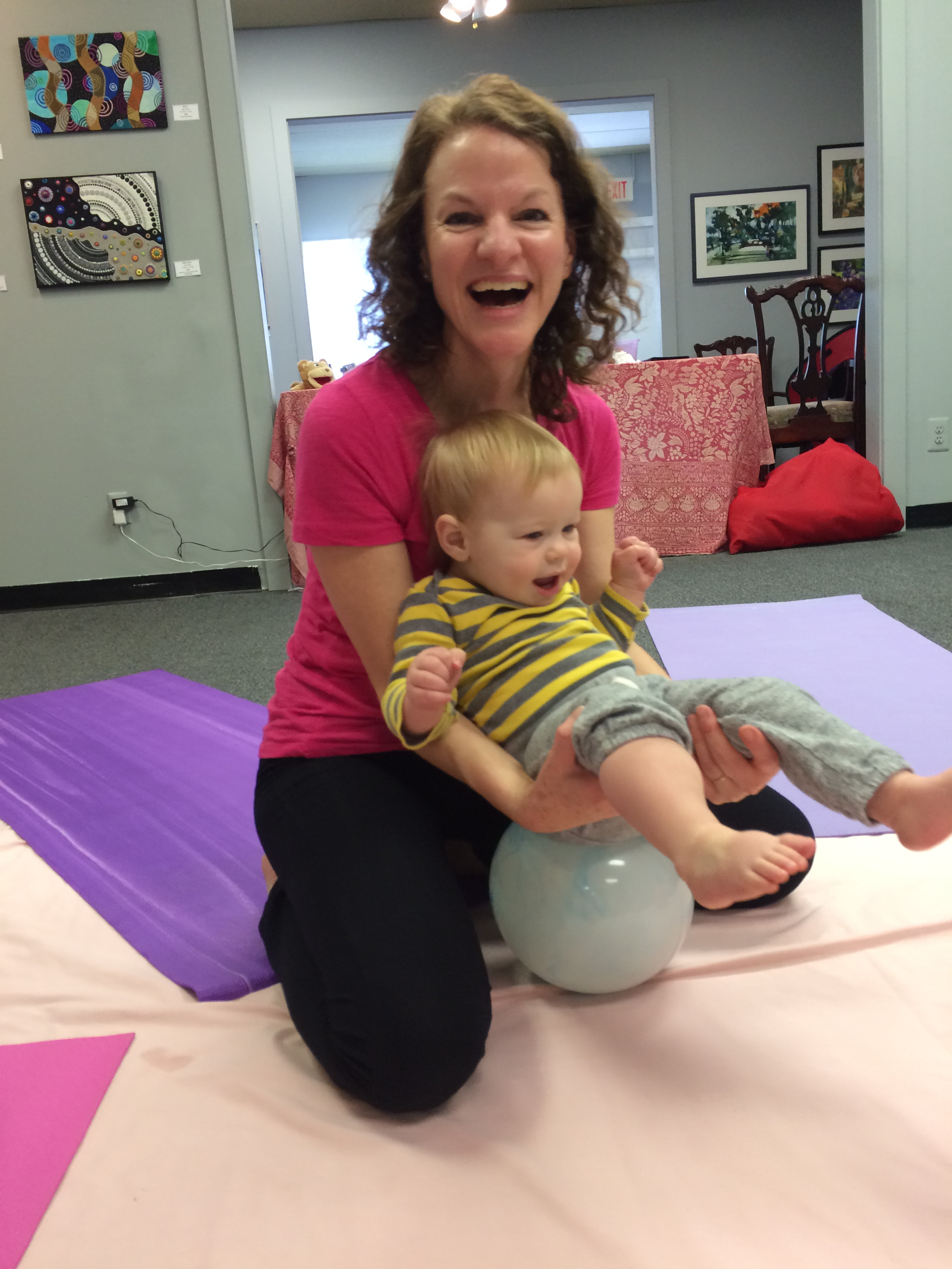 A woman with curly brown hair is sitting on her knees on a pink mat, laughing, and holding a toddler with blonde hair and a striped yellow and gray shirt, who is also smiling and sitting on a white exercise ball.