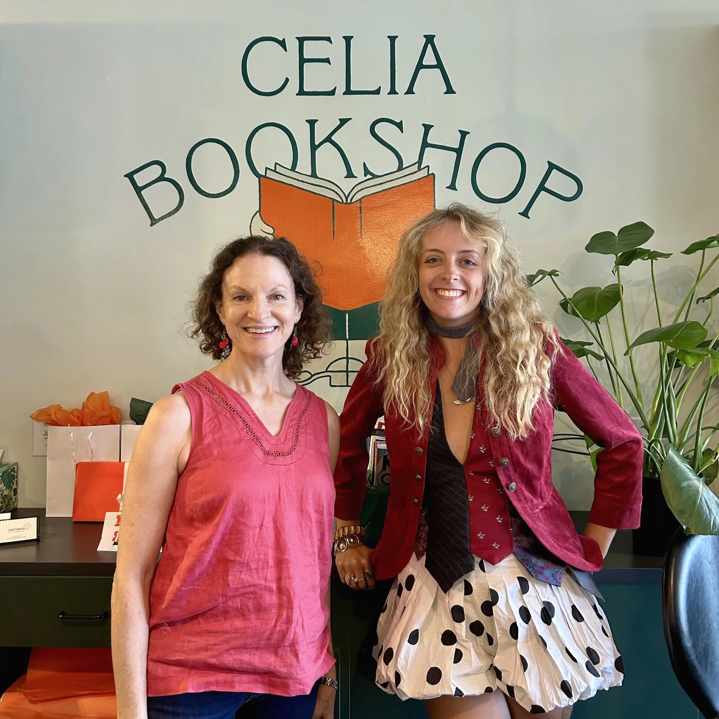 Two women smiling and standing in front of a wall with a sign that says 'Celia Bookshop' and an illustration of an open book. There are green plants in the foreground.