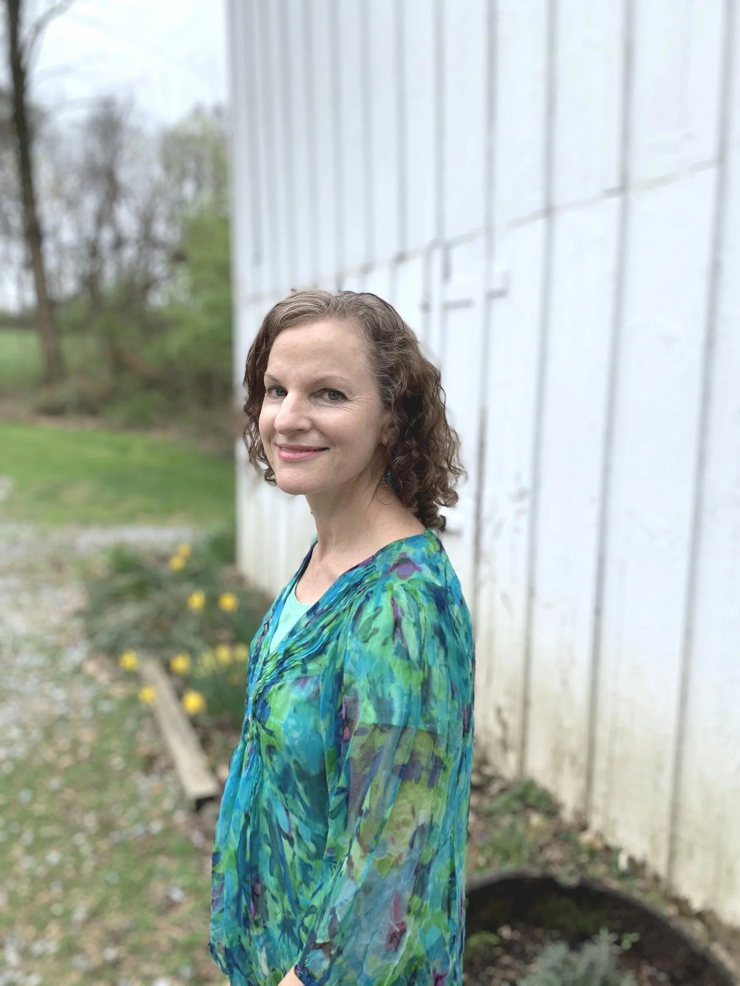 A woman with curly brown hair smiling while standing outdoors next to a white fence and a tree with green leaves in the background.