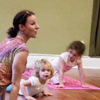 A woman on a pink yoga mat with two young girls practicing yoga in a room with light green walls.