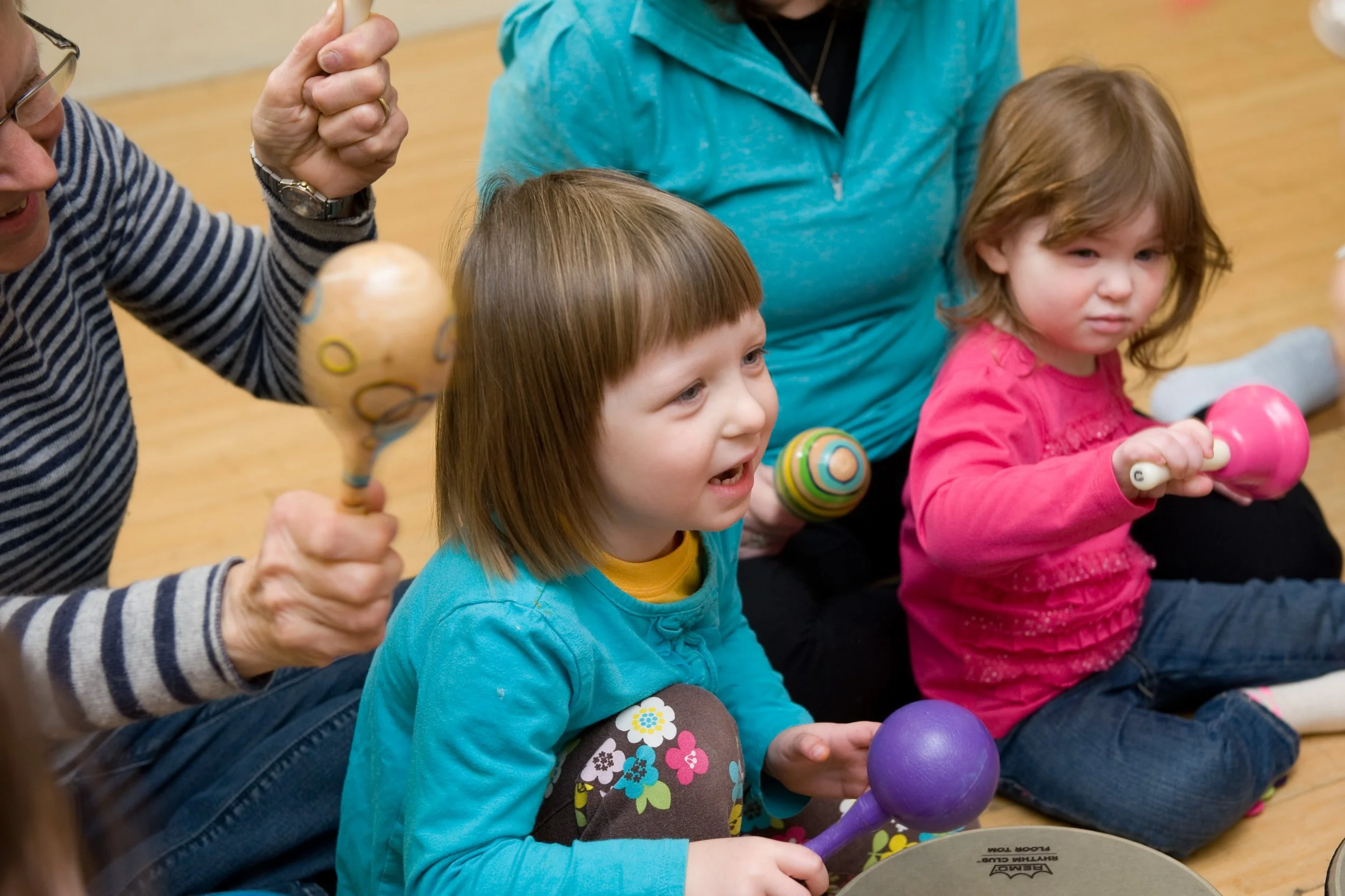 Children sitting on the floor at a music class, holding percussion instruments like maracas and shakers, with a few adults assisting.