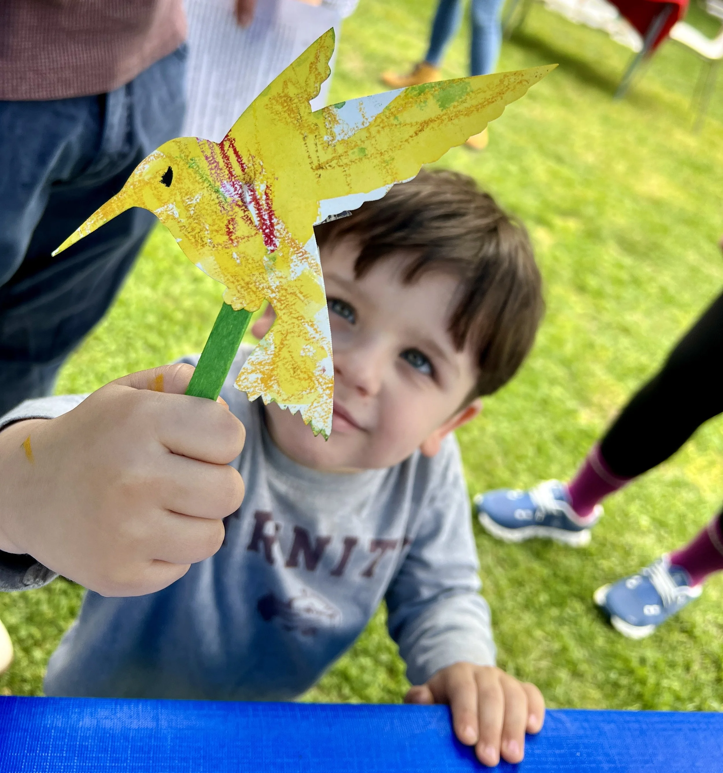 Child holding a colorful paper hummingbird craft outdoors on green grass.
