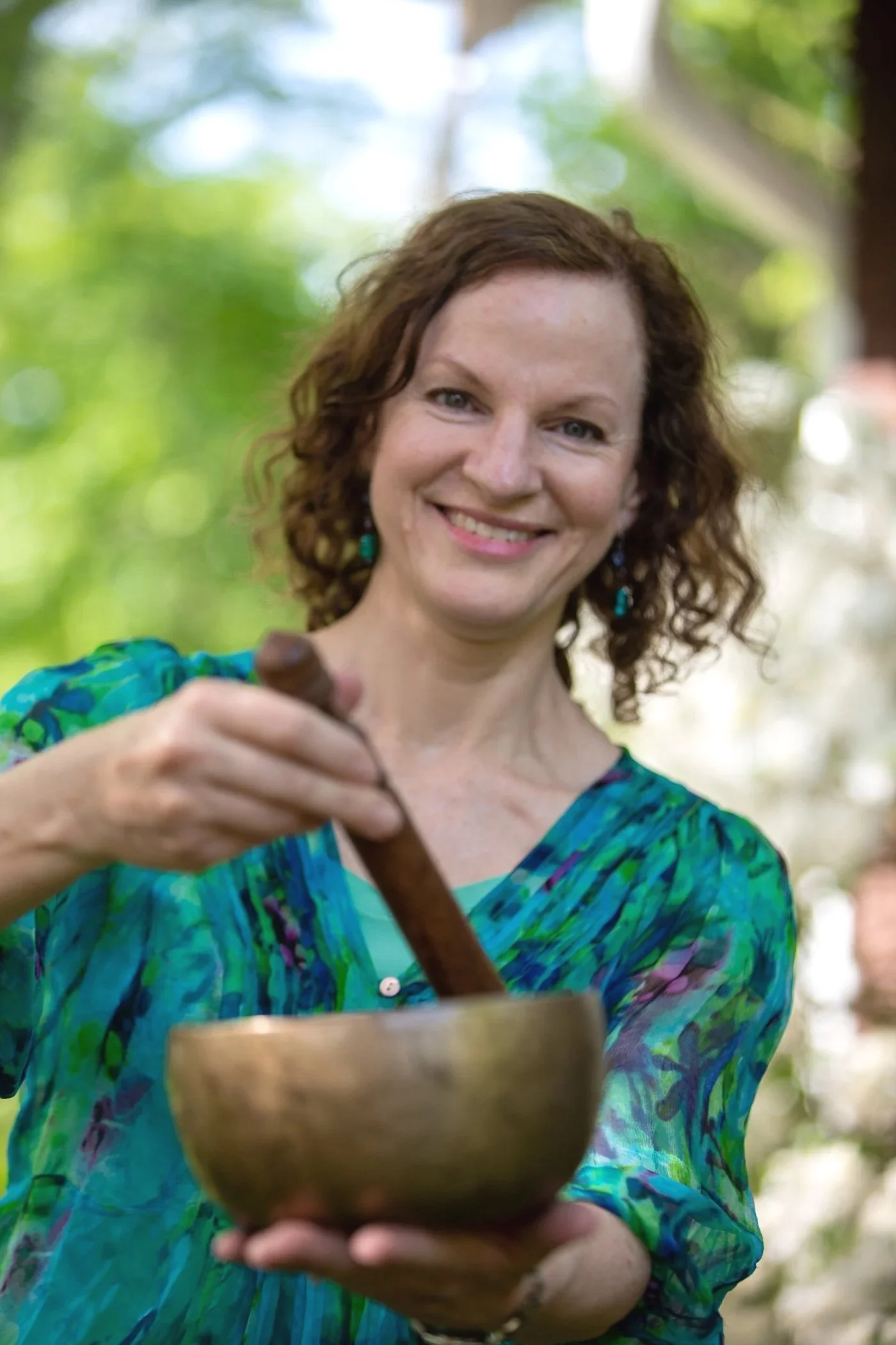 A woman with curly red hair and blue earrings smiling and holding a wooden spoon over a bowl outdoors with green trees in the background.