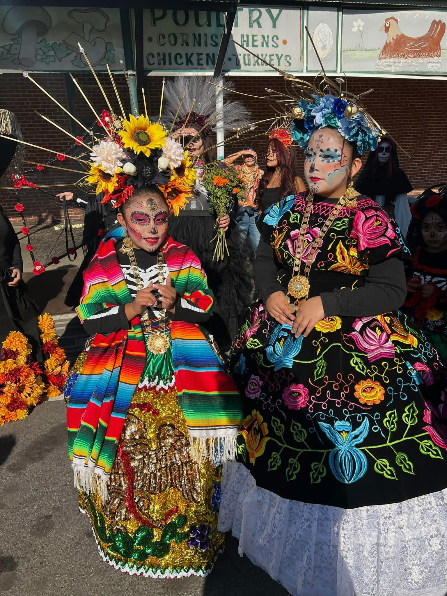 Two women dressed in traditional Mexican attire with colorful embroidered dresses, elaborate floral and feather headpieces, and face paint resembling sugar skulls, standing at a festive event.