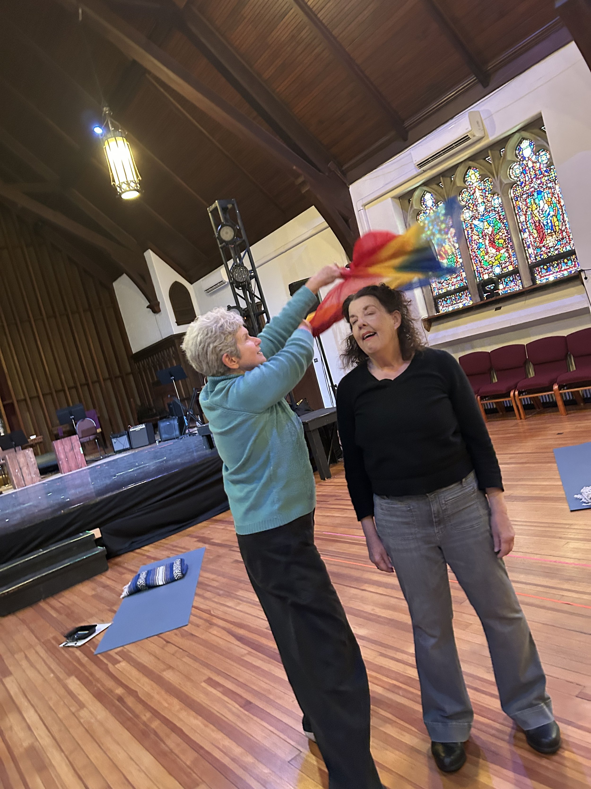 Two women are inside a room with a wooden ceiling and stained glass windows. One woman is holding a rainbow-colored cloth over the other woman's head. The room has musical equipment and chairs in the background.