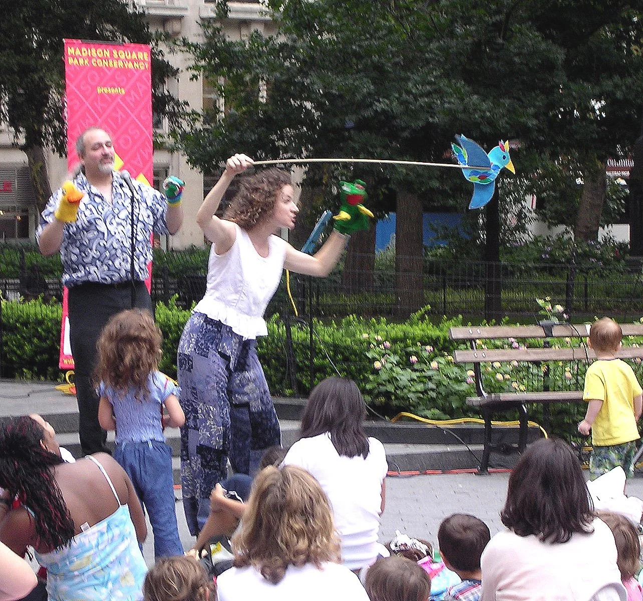 A young woman performing a puppet show with a blue bird puppet in a park, with children and adults sitting and watching.