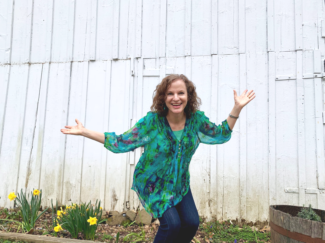 Woman smiling and gesturing outside near a white wooden fence and yellow flowers.