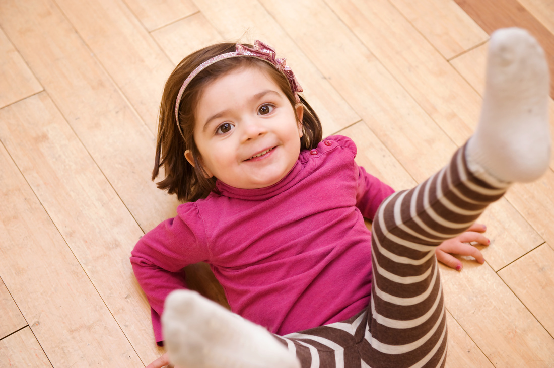 Cute young girl with shoulder-length brown hair, wearing a pink long-sleeve top and striped leggings, smiling and lying on a wooden floor with one leg raised.