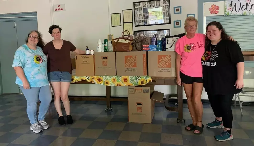 Four women standing in front of a table with boxes and supplies, inside a room with framed pictures and a 'No Smoking' sign on the wall.