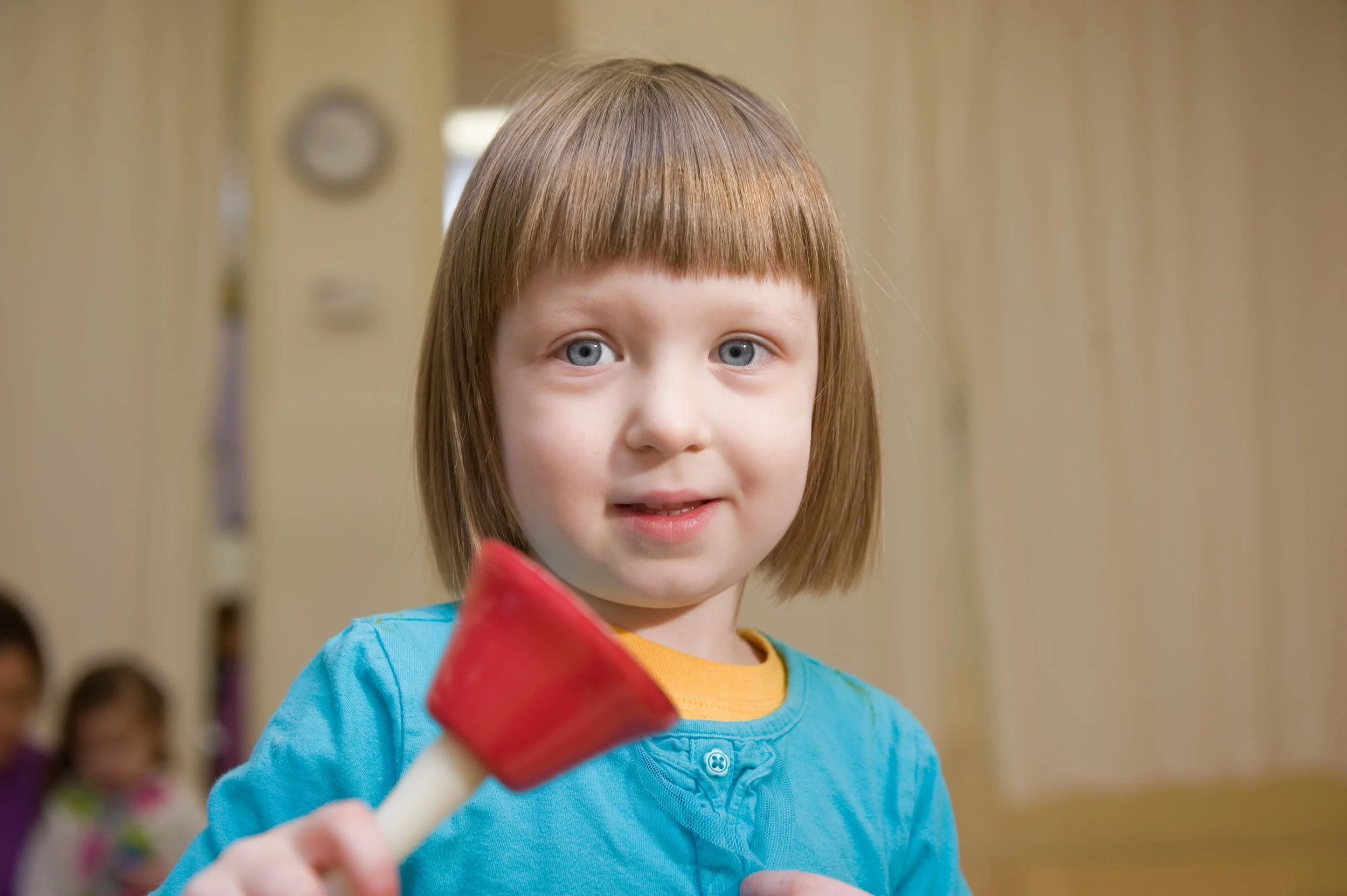 A young girl with brown hair and blue eyes holding a red and white object, wearing a blue shirt, in a classroom with other children blurred in the background.