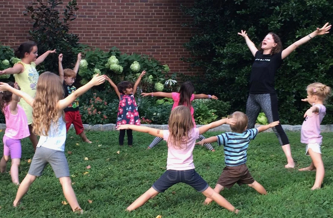 Children playing in a circle on the grass with an adult leading. All are raising their arms and smiling, enjoying outdoor activity.