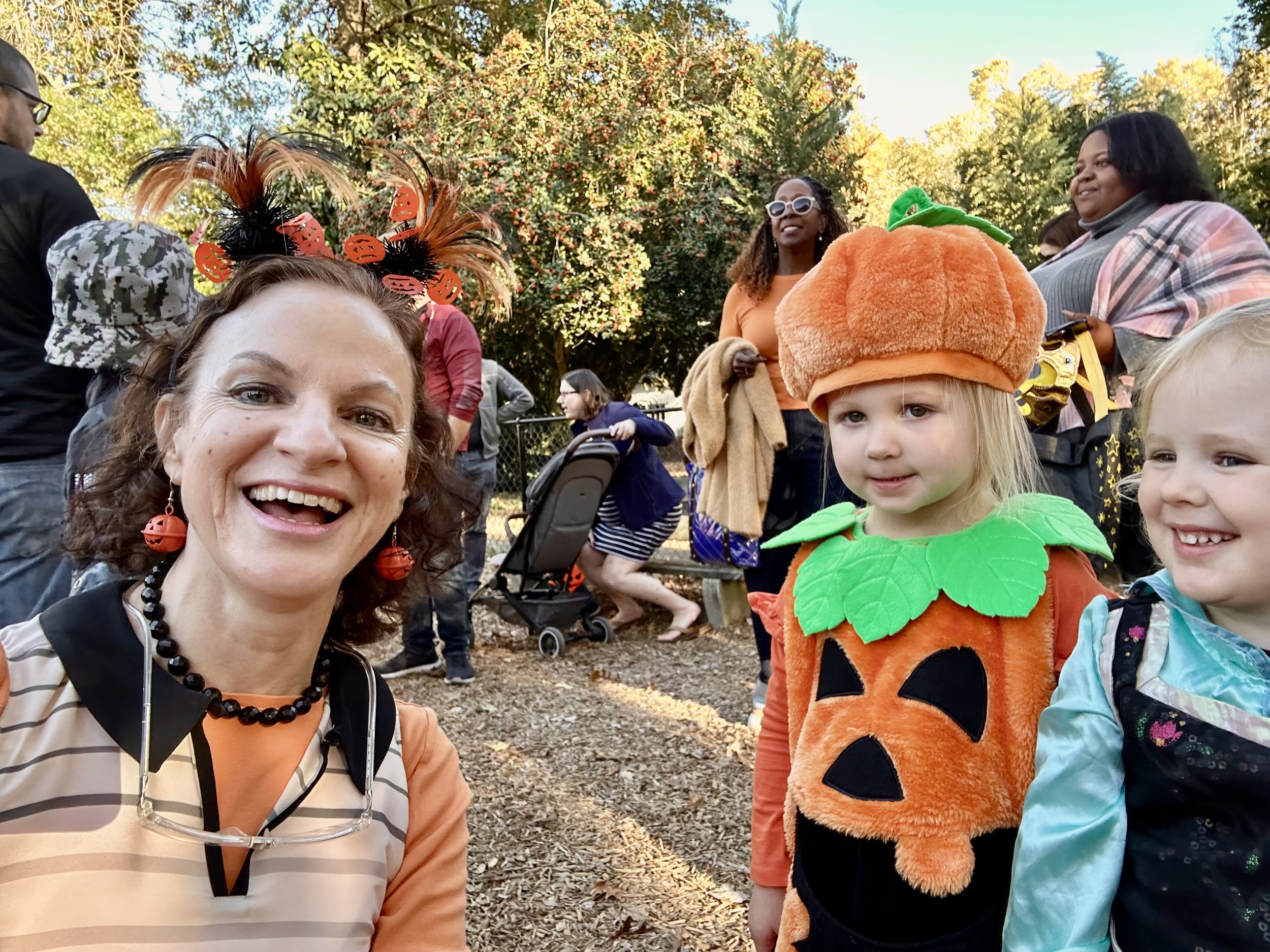 Group of people celebrating Halloween outdoors, with children in costumes including a pumpkin and a dragon, and adults smiling.