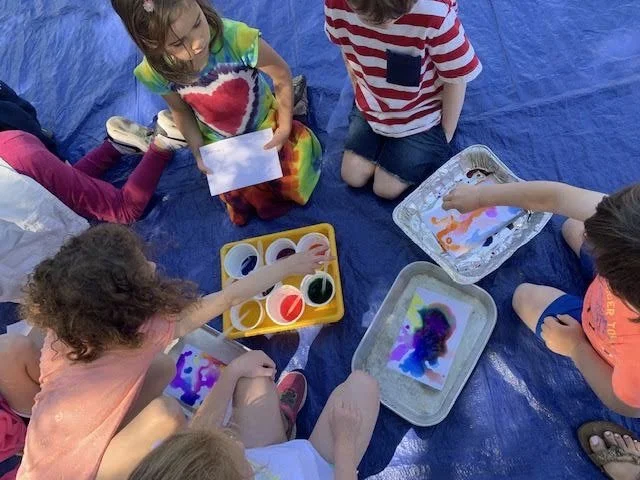 Children sitting on a blue tarp, engaging in a watercolor painting activity with various paint colors and brushes.