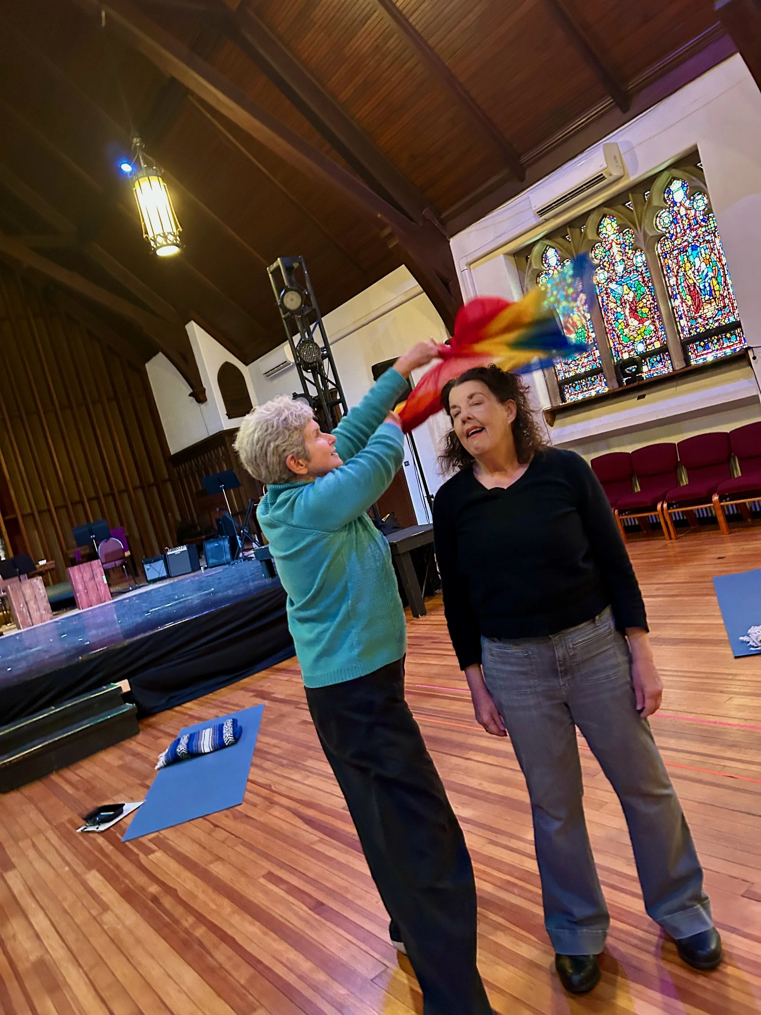 Two women in a church hall, one holding a rainbow-colored fabric above the other’s head. The hall has a wooden ceiling, stained glass windows, and a stage with audio equipment.