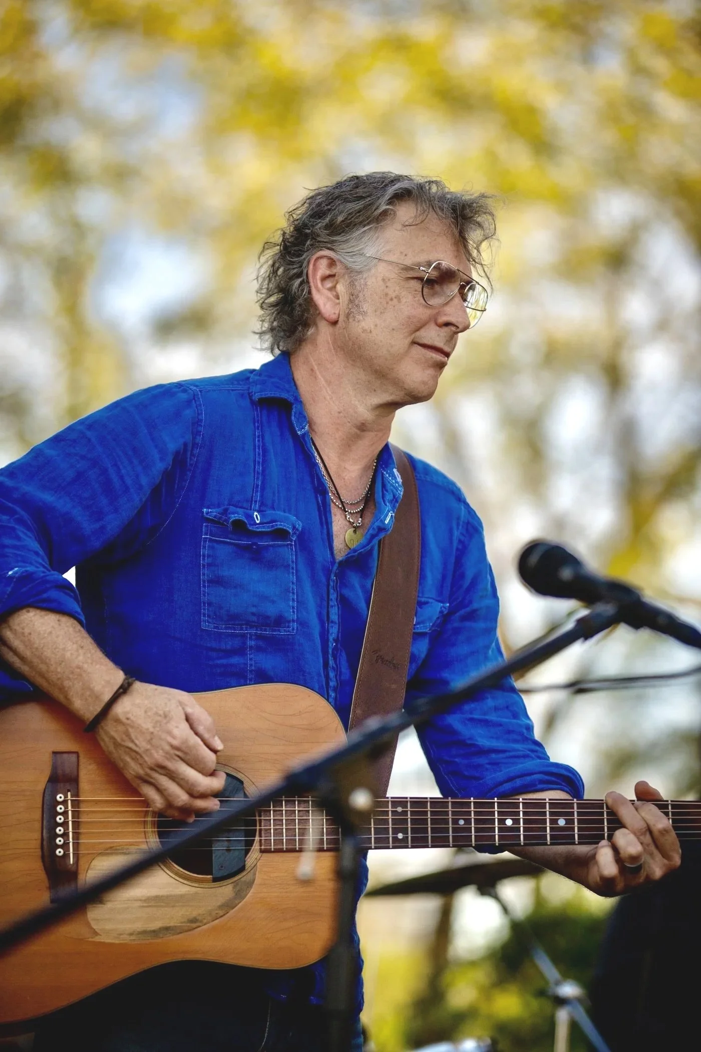 A man with gray, curly hair, wearing glasses and a blue shirt, is playing an acoustic guitar outdoors with a blurred background of trees with yellow leaves.