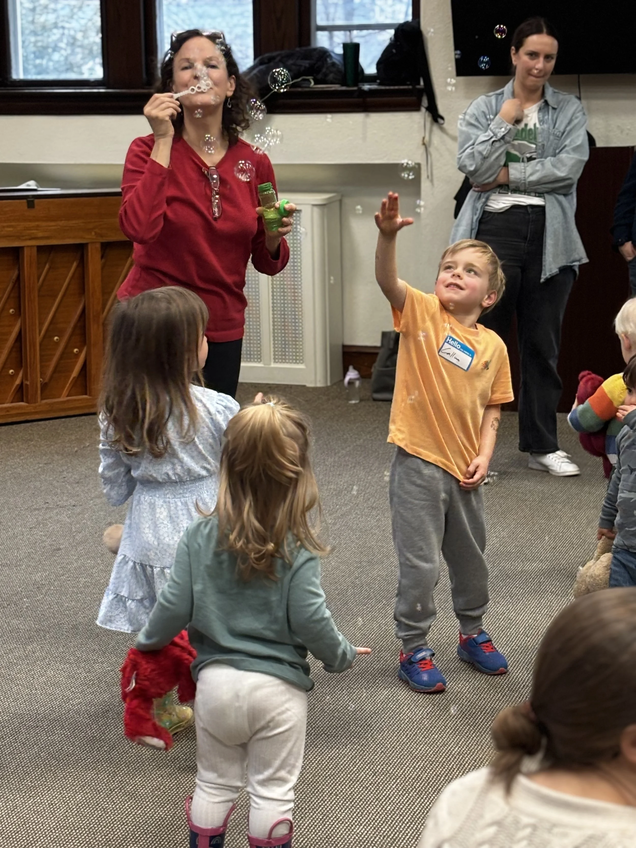 Children and adults playing and watching bubbles in a room with carpeted floor and windows.