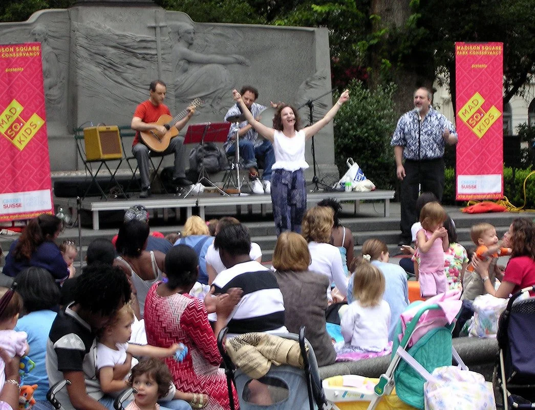 Children and families watching a live outdoor performance with musicians and a woman singing on a stage in front of a monument.