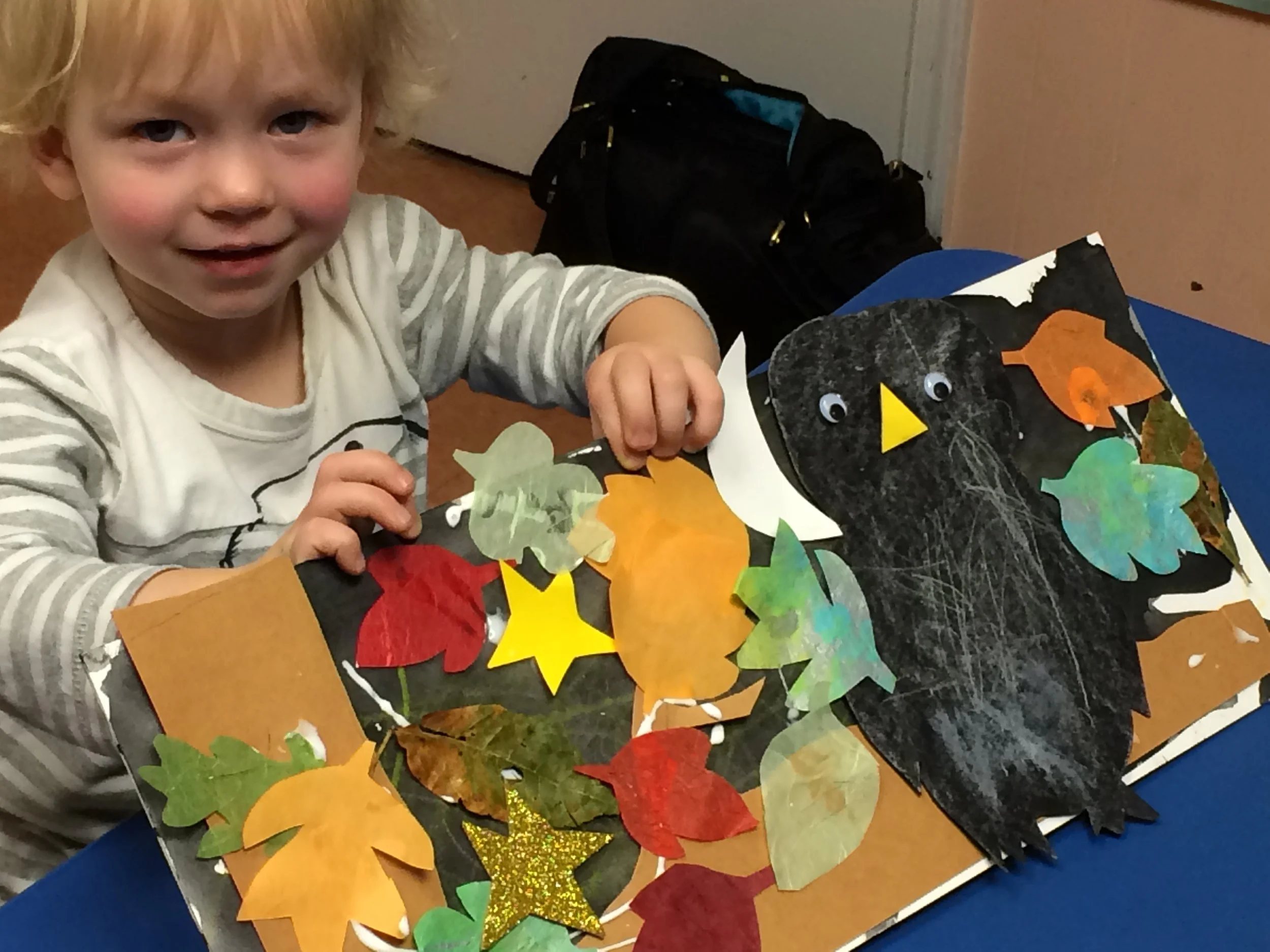 A young boy with blonde hair smiling at a table decorated with colorful paper cutouts of fall leaves and a black turkey craft with googly eyes and an orange beak.