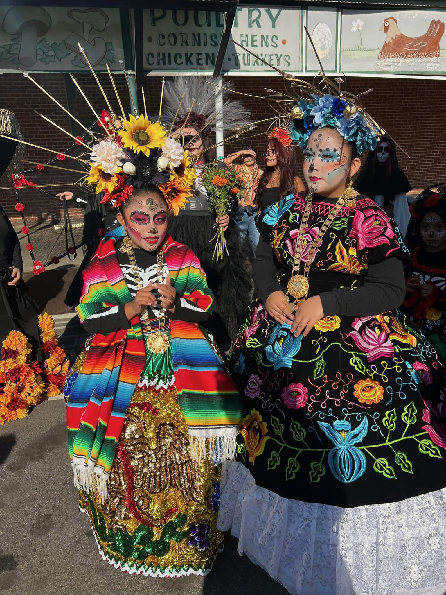 Two women dressed in colorful traditional Mexican attire with intricate face paint and large floral headpieces, standing in front of a market stall.