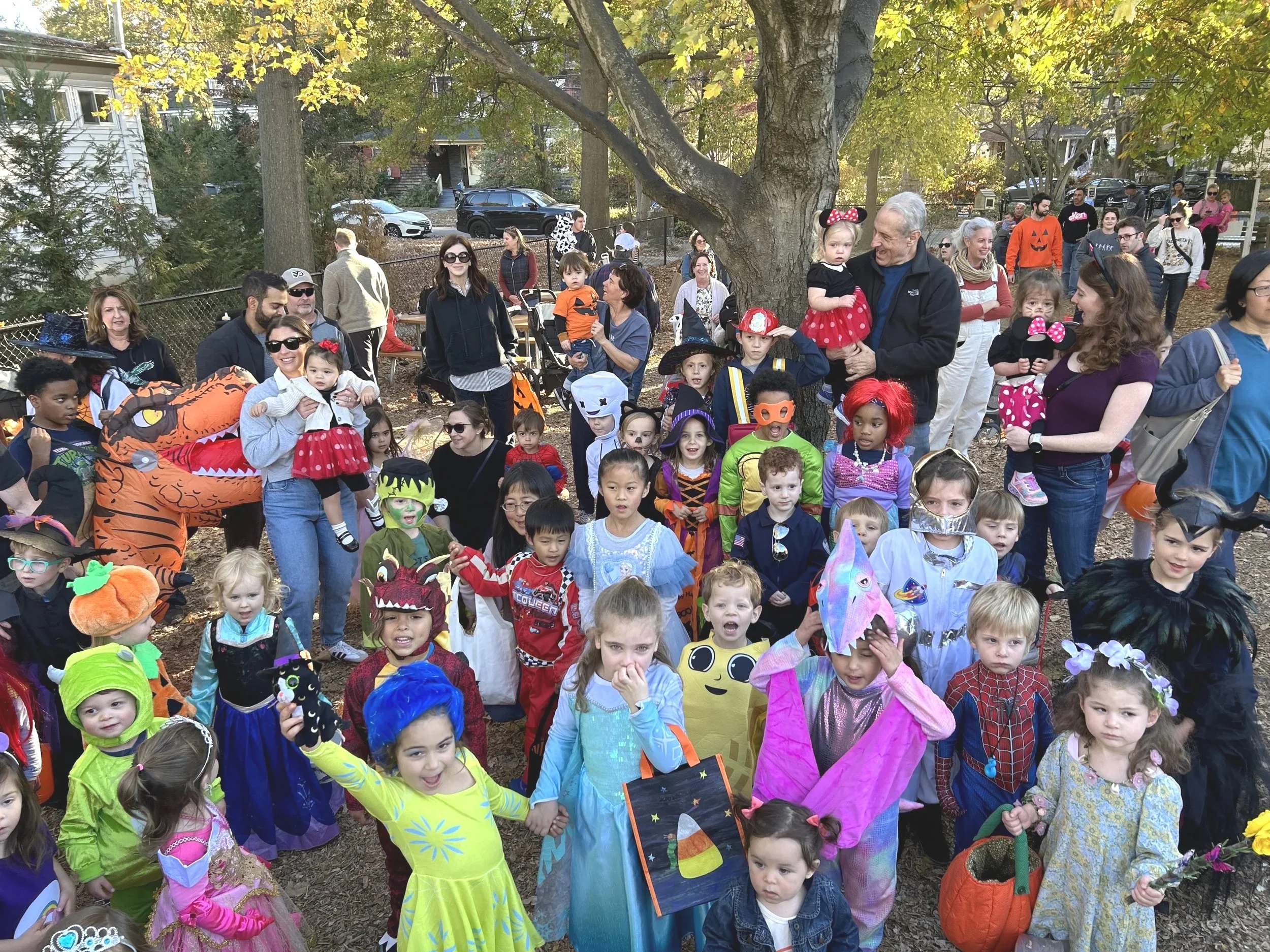 Group of children and adults dressed in costumes for a Halloween or costume party in a park with trees and cars in the background.
