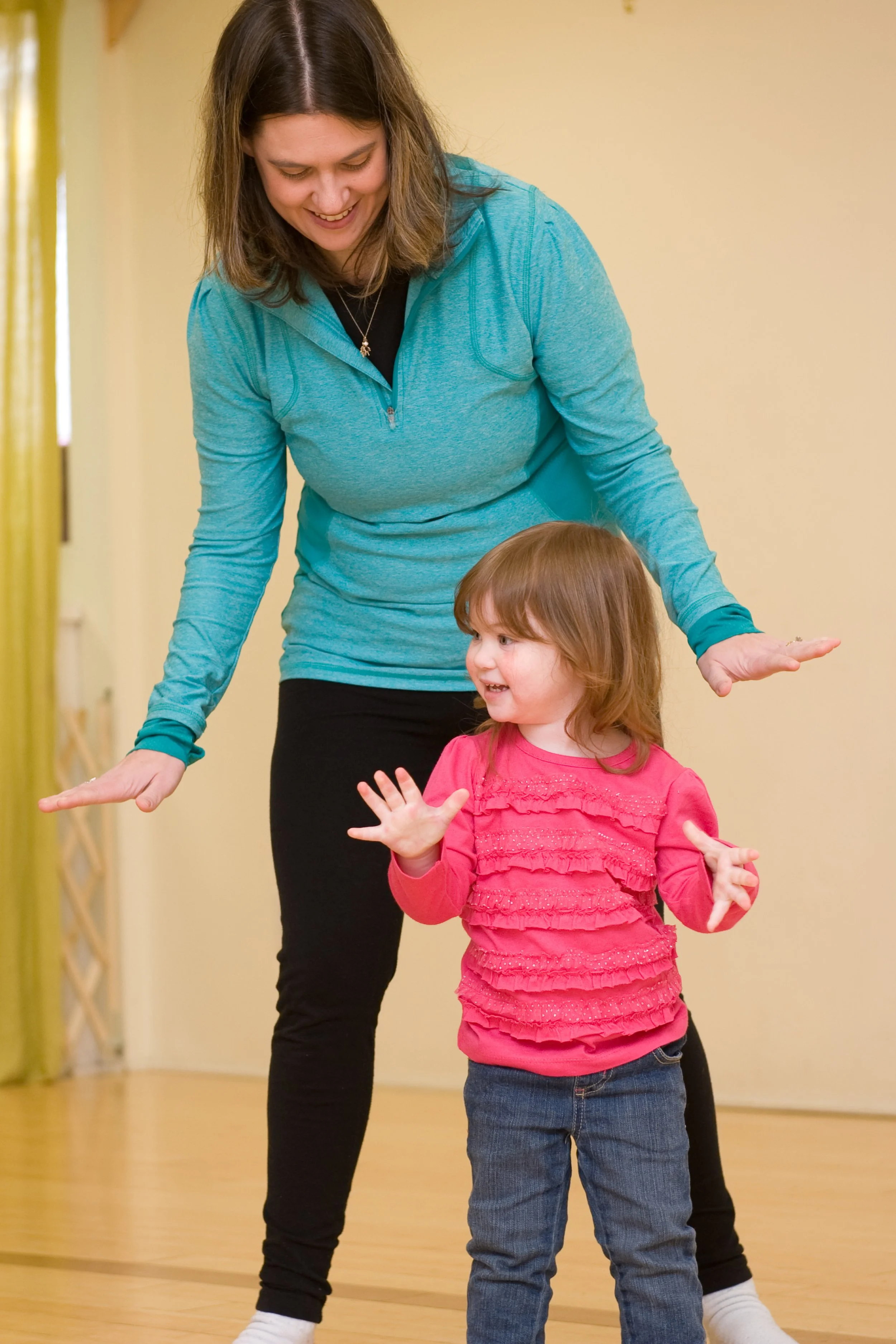 A woman and a young girl are practicing dance or movement, with the woman bending over slightly and the girl mid-motion, both smiling and engaging with hand gestures. The woman has shoulder-length brown hair, wears a teal long-sleeve top and black pants, and the girl has shoulder-length brown hair, a pink ruffled top, and jeans. They are in an indoor space with yellow walls and a wooden floor.