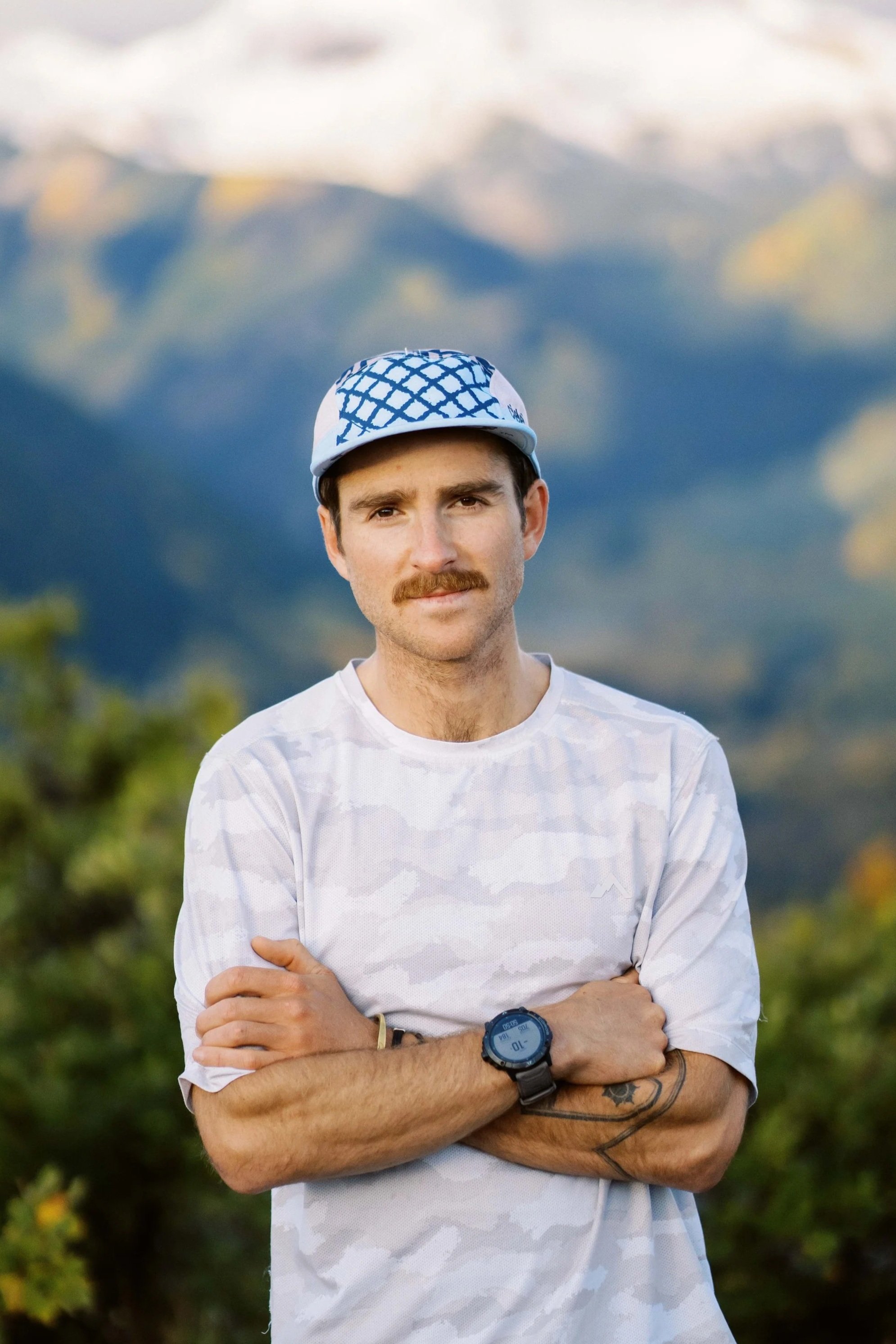 A man with a mustache, wearing a white shirt, a watch, and a cap with a geometric pattern, standing outdoors with hills or mountains in the background.