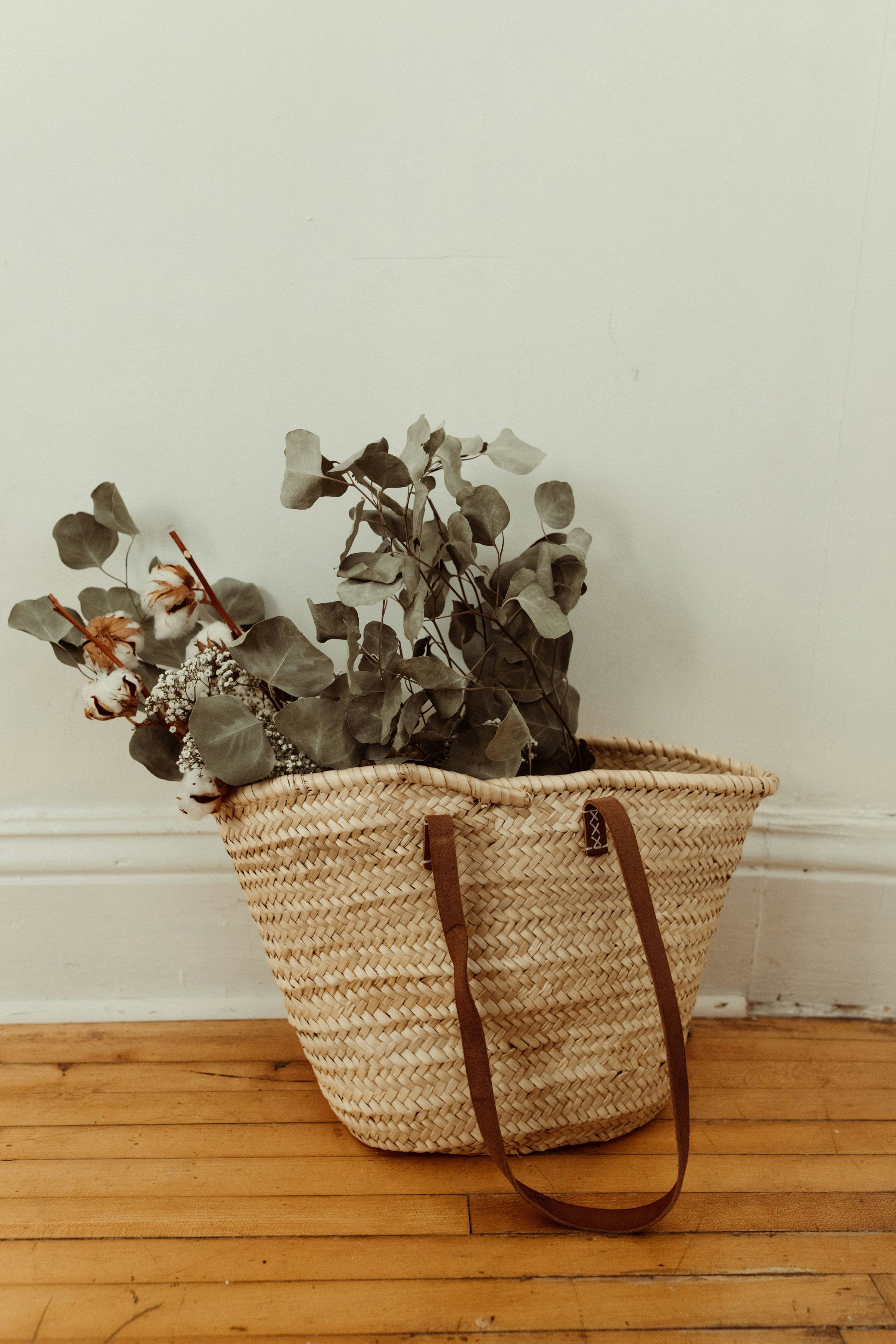 A woven basket with leather straps sitting on a hardwood floor against a plain wall, holding dried eucalyptus and cotton flowers.